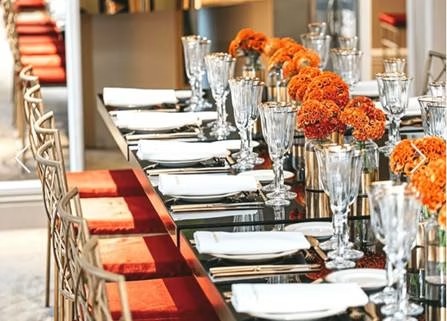 Mesa larga con copas altas, vajilla blanca y flores naranjas